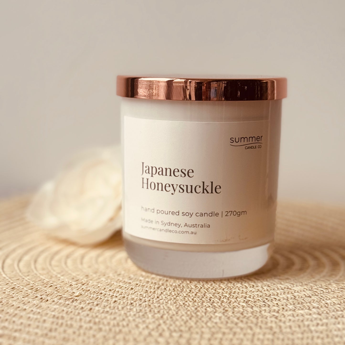 Hand-poured soy wax candle in a white jar with a rose gold lid, scented with Japanese Honeysuckle. A sweet floral aroma with notes of jasmine and honey. Clean-burning and perfect for gifting or creating a calming, elegant atmosphere.