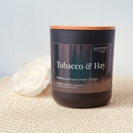 Hand-poured soy wax candle in a sleek black jar with a natural wooden lid, scented with Tobacco & Hay. Features earthy notes of dried hay, tobacco leaf, and musk. A clean-burning, masculine fragrance ideal for creating a warm, rustic atmosphere.