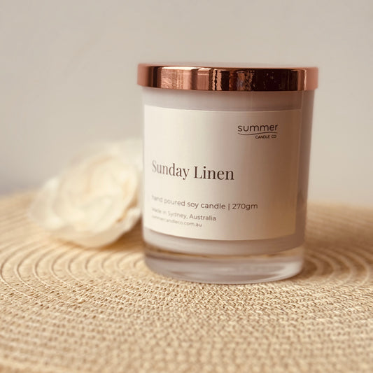 Clear glass soy wax candle jar labeled “Sunday Linen” with a sleek rose gold lid, featuring minimalist and elegant packaging. The candle evokes fresh, clean linen scents, perfect for creating a calm and cozy atmosphere at home. Ideal as a stylish gift or for enhancing your living space with subtle, soothing fragrance and modern home decor appeal.